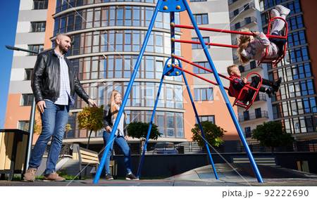Happy family - father, mother and children having fun together on playground in new city district. Parents swinging daughter and son. Modern residential buildings on background. 92222690
