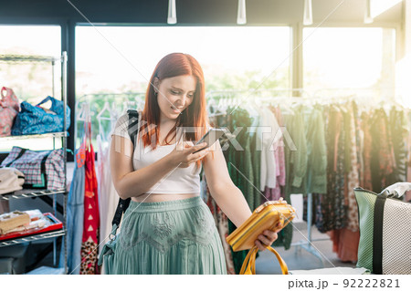 young red-haired woman happy shopping in a clothing shop, taking pictures with her smartphone of the clothes. girl spending time shopping sharing it on her social networks. concept of shopping. 92222821
