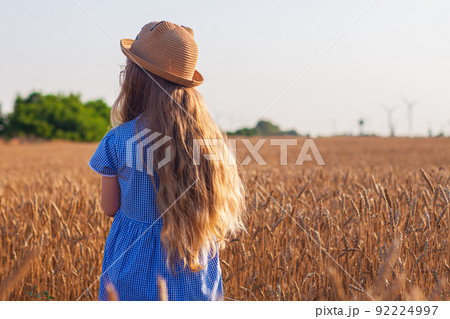 Adorable little girl in a straw hat, blue plaid summer dress in wheat field countryside.Child with long blonde wavy hair 92224997