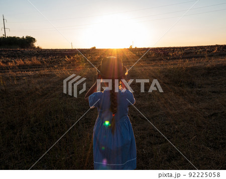 Adorable little girl in a straw hat, blue plaid summer dress in grass field countryside. Child with long blonde braid. 92225058