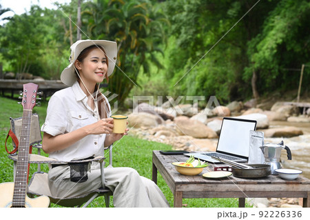 Happy female traveler drinking coffee, resting in folding chair near the river bank. Adventure, travel and camping concept 92226336
