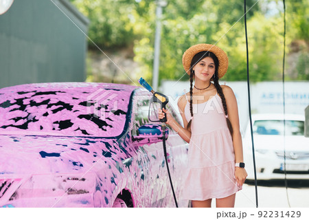 Woman with hose stands by car covered in pink foam 92231429