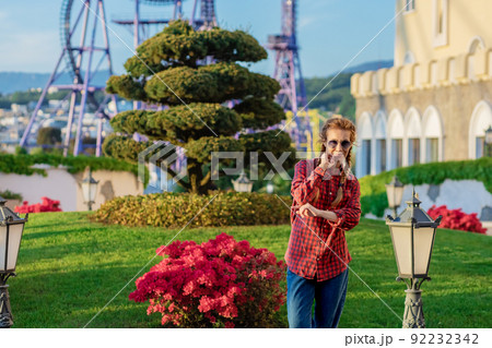 A woman in a youth shirt and jeans poses standing by a flower bed against the backdrop of an amusement park and a cityscape in the sunset 92232342