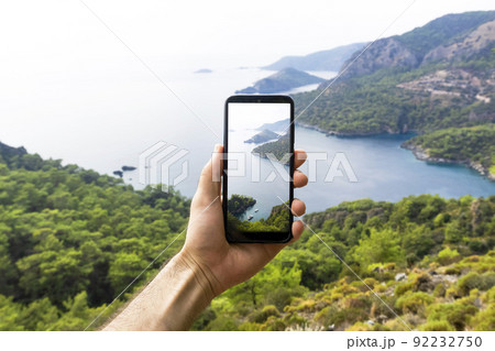 A hand with a smartphone taking pictures of the natural landscape, sea and mountains on a journey. Phone, internet and mobile network concept. Impressions and Adventures. 92232750