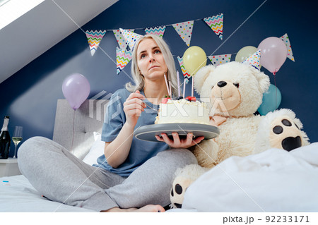 Sad, upset woman in pajama and party cap eating by spoon her birthday cake sitting on the bed with huge teddy bear in decorated bedroom. Celebrates birthday alone. Jamming of negative emotions. Sad, upset woman in pajama and party cap eating by spoon her birthday cake sitting on the bed with huge teddy bear in decorated bedroom. Celebrates birthday alone. Jamming of negative emotions. 92233171