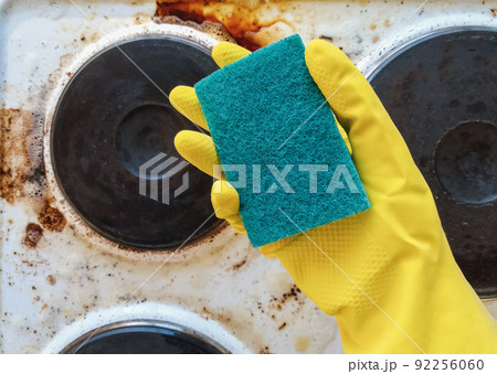 hand in yellow glove holding washup sponge against dirty electric stove 92256060