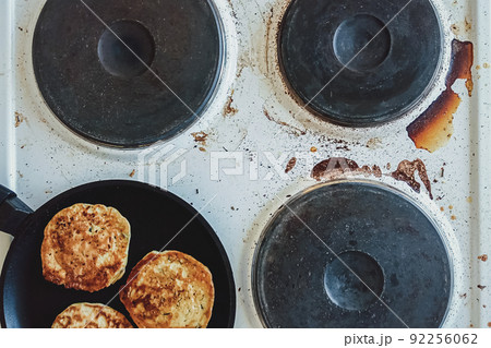 kitchen stove dirty with burnt food and cooking oil, frying pan with pancakes on top 92256062