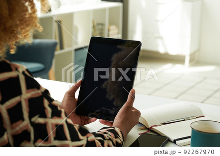 Close-up of African girl looking at screen of digital tablet and working online sitting at table with notepad in the room Close-up of African girl looking at screen of digital tablet and working online sitting at table with notepad in the room 92267970