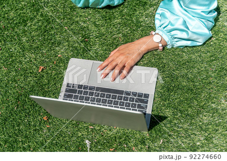 a young beautiful woman with blond curly hair in glasses and a blue dress sits on the grass in nature and uses a laptop, 92274660