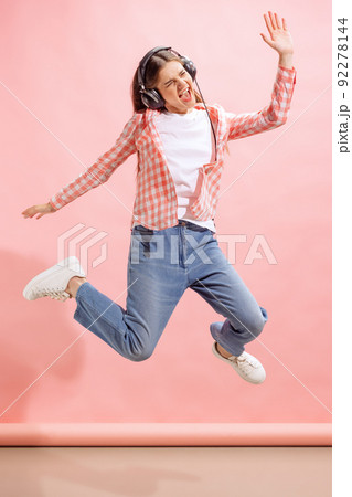 Portrait of emotive young girl in checkered shirt listening to music in headphones and jumping isolated over pink studio background 92278144