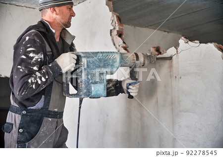 Man builder in workwear drilling wall with electric hammer drill. Male worker using electric drill while breaking wall in apartment under renovation. Demolishing and construction destroying concept. Man builder in workwear drilling wall with electric hammer drill. Male worker using electric drill while breaking wall in apartment under renovation. Demolishing and construction destroying concept. 92278545