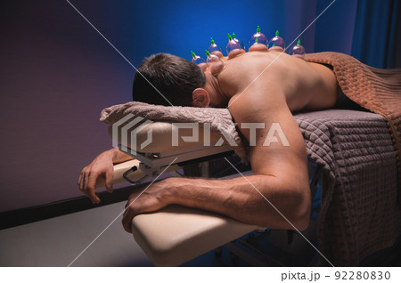 Relaxed young muscular man getting cupping treatment on his back lying on table Relaxed young muscular man getting cupping treatment on his back lying on table 92280830