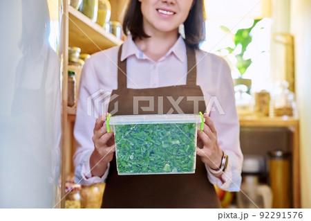 Woman with container of frozen green onions, in kitchen in pantry near freezer. 92291576