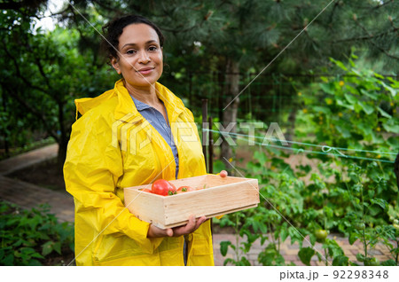 Inspired cheerful successful woman gardener in a yellow raincoat in allotment garden, smiling looking at the camera. 92298348