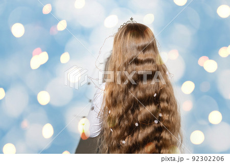 Rear view of a girl with long wavy flowing hair on a bright shimmering blue and yellow background. A crown and garlands of snowflakes for a child. Merry Christmas and Happy New Year. Copy space. 92302206