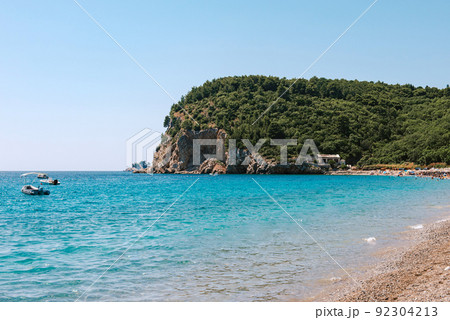 Picturesque view on rocks on a sunny day from the sea. Budva riviera, Montenegro. Aerial view of Sveti Nikola, Budva island, Montenegro. Hawaii beach, umbrellas and bathers and crystal clear waters 92304213