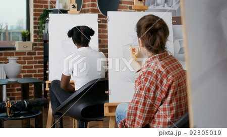 Young woman using pencil to draw vase on white canvas, learning new artistic skills in art class. Student drawing inspiration model at lesson practice with multi ethnic group of people. 92305019