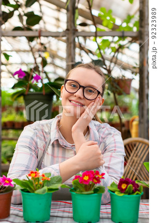 Vertical photo of a young woman with a few flowers in a greenhouse - she looking at camera and smiling. 92323989