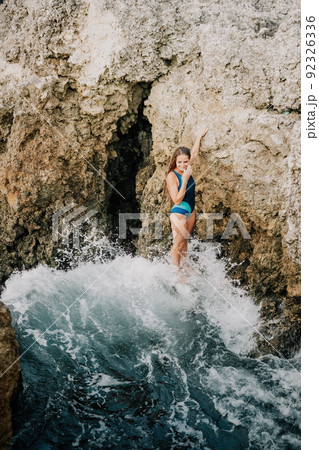 A young beautiful tanned woman with long hair in blue bikini sitting at rocks near blue green clear ocean sea water. Woman relax on sunset at sea, enjoy holidays and weekend vacation in summer time. 92326336