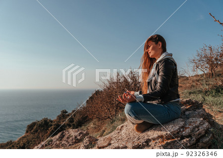 A young tourist Woman enjoying sunset over sea mountain landscape while sitting outdoor. Women's yoga fitness routine. Healthy lifestyle, harmony and meditation 92326346