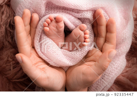 The palms of the parents. A father and mother hold the feet of a newborn child in a pink blanket on a brown background.. The feet of a newborn in the hands of parents. Photo of foot, heels and toes The palms of the parents. A father and mother hold the feet of a newborn child in a pink blanket on a brown background.. The feet of a newborn in the hands of parents. Photo of foot, heels and toes 92335447