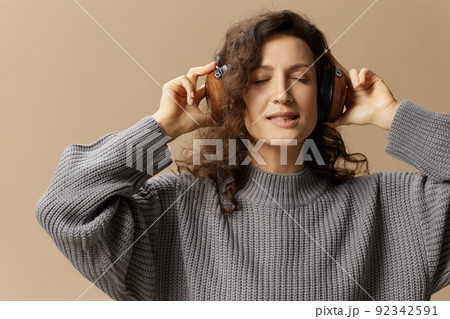 Pretty enjoyed curly beautiful female in gray casual sweater headphones listen favorite song posing isolated on over beige pastel background. Sound streaming platform concept. Copy space Pretty enjoyed curly beautiful female in gray casual sweater headphones listen favorite song posing isolated on over beige pastel background. Sound streaming platform concept. Copy space 92342591