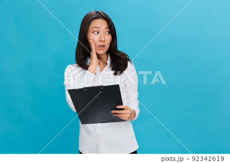 Shocked cute Asian businesswoman in classic office dress code holds folder tablet tells secret posing isolated on over blue studio background. Cool business offer. Job interview concept 92342619