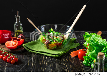 A glass bowl of fresh vegetable salad surrounded by the ingredients: green lettuce, cucumbers, sweet pepper and tomatoes. A glass bowl of fresh vegetable salad surrounded by the ingredients: green lettuce, cucumbers, sweet pepper and tomatoes. 92348702