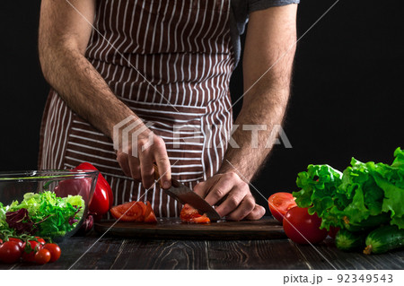 Man preparing salad on a wooden table. Men's hands cut the tomato to make a salad on black background Man preparing salad on a wooden table. Men's hands cut the tomato to make a salad on black background 92349543