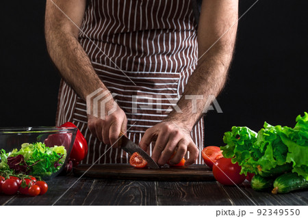 Man preparing salad on a wooden table. Men's hands cut the tomato to make a salad on black background Man preparing salad on a wooden table. Men's hands cut the tomato to make a salad on black background 92349550
