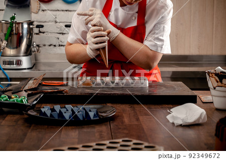 A woman confectioner with red uniform and white sterile gloves do a set of colorful chocolates from milk chocolate on a table. A woman confectioner with red uniform and white sterile gloves do a set of colorful chocolates from milk chocolate on a table. 92349672