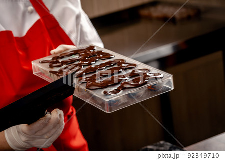 A woman confectioner with red uniform and white sterile gloves do a set of colorful chocolates from milk chocolate on a table. A woman confectioner with red uniform and white sterile gloves do a set of colorful chocolates from milk chocolate on a table. 92349710
