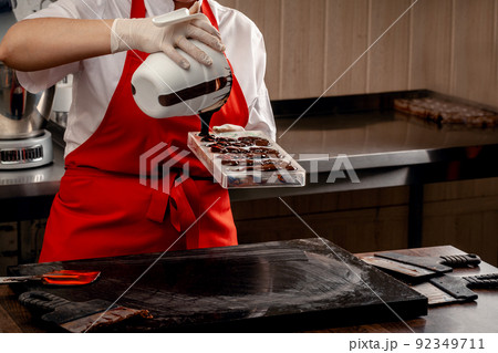 A woman confectioner with red uniform and white sterile gloves do a set of colorful chocolates from milk chocolate on a table. A woman confectioner with red uniform and white sterile gloves do a set of colorful chocolates from milk chocolate on a table. 92349711
