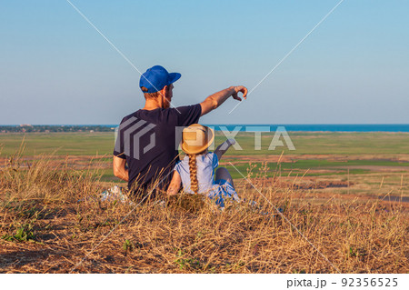 Father and daughter sitting by lakes on a nature lanscape. A little girl with long blond hair braid hugging dad outdoor. 92356525