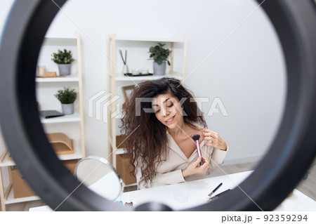 Professional makeup artist tanned handsome curly Latin beauty blogger in linen shirt tells how to sculpt body in home white interior. Copy space. Influencer record video review blog using smartphone Professional makeup artist tanned handsome curly Latin beauty blogger in linen shirt tells how to sculpt body in home white interior. Copy space. Influencer record video review blog using smartphone 92359294