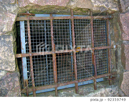 A grate covering the entrance to a stone dungeon, a cave. An old metal forged grate closes a hole in the rock A grate covering the entrance to a stone dungeon, a cave. An old metal forged grate closes a hole in the rock 92359749