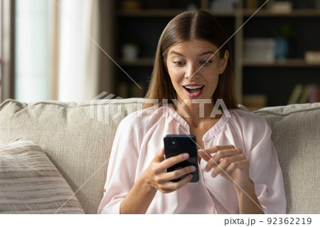 Woman holds smartphone staring at screen read sms looks stunned Woman holds smartphone staring at screen read sms looks stunned 92362219