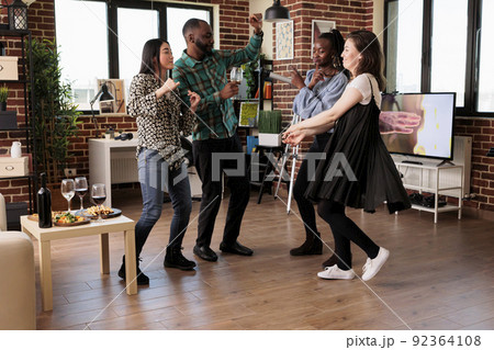 Cheerful friends having celebration event for friendship anniversary while drinking alcoholic beverages. Diverse group of young people dancing in living room at wine party while celebrating birthday. Cheerful friends having celebration event for friendship anniversary while drinking alcoholic beverages. Diverse group of young people dancing in living room at wine party while celebrating birthday. 92364108