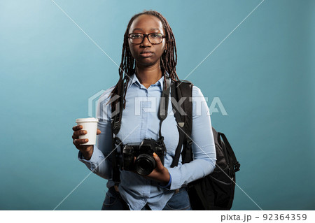 Serious looking professional photographer having DSLR device and cup of coffee while looking at camera on blue background. Young photography entusiast having photo equipment while enjoying beverage. 92364359