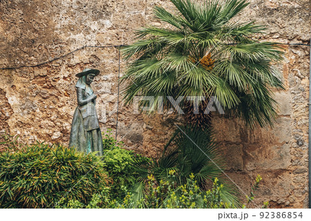 Female statue by palm tree amidst plants against wall in town 92386854