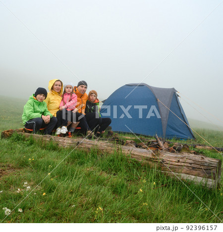 A family in bright clothes sits near a tent in the mountains, morning fog in the mountains. 92396157
