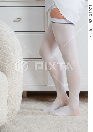 White hosiery. Beautiful long female legs in stockings. Girl putting on stockings at home in a white room. Varicose veins prevention. Woman body in underwear. 92404075
