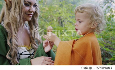 Fairy elf cosplay woman treats little baby boy hobbit with candy in green forest. Halloween concept 92409803
