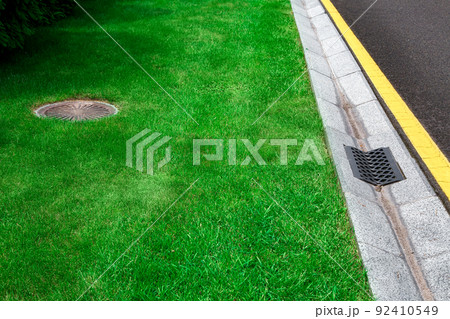 gray gutter of a stormwater system on the side of asphalt road with yellow markings and manhole in green trimmed lawn, concrete drainage ditch with iron grate on sunny dry weather, nobody. gray gutter of a stormwater system on the side of asphalt road with yellow markings and manhole in green trimmed lawn, concrete drainage ditch with iron grate on sunny dry weather, nobody. 92410549
