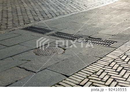 round iron manhole with hatches rectangular gratings of the drainage system on the road with paved stone block near the pedestrian sidewalk made of brick tile close-up view with sun light, nobody. round iron manhole with hatches rectangular gratings of the drainage system on the road with paved stone block near the pedestrian sidewalk made of brick tile close-up view with sun light, nobody. 92410555