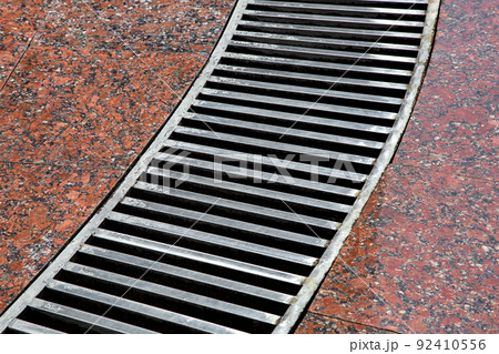 curved drainage system with stripes grate on the granite floor with a wet red stone granite surface, close-up of a storm system wet after rain weather, nobody. curved drainage system with stripes grate on the granite floor with a wet red stone granite surface, close-up of a storm system wet after rain weather, nobody. 92410556