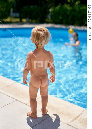 Small naked baby stands by the pool with turquoise water. Back view 92417197