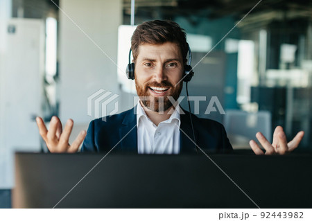 Happy handsome businessman in headset working on laptop and gesturing to webcam, having video call in office interior Happy handsome businessman in headset working on laptop and gesturing to webcam, having video call in office interior 92443982