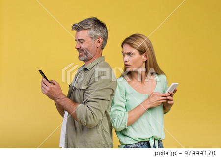 Shocked woman spying on her happy husband using smartphone, standing back to back over yellow background Shocked woman spying on her happy husband using smartphone, standing back to back over yellow background 92444170