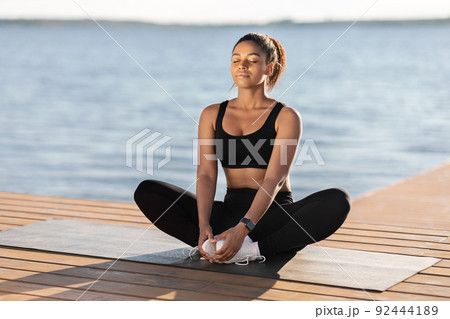 Peaceful young black woman meditating next to city lake 92444189